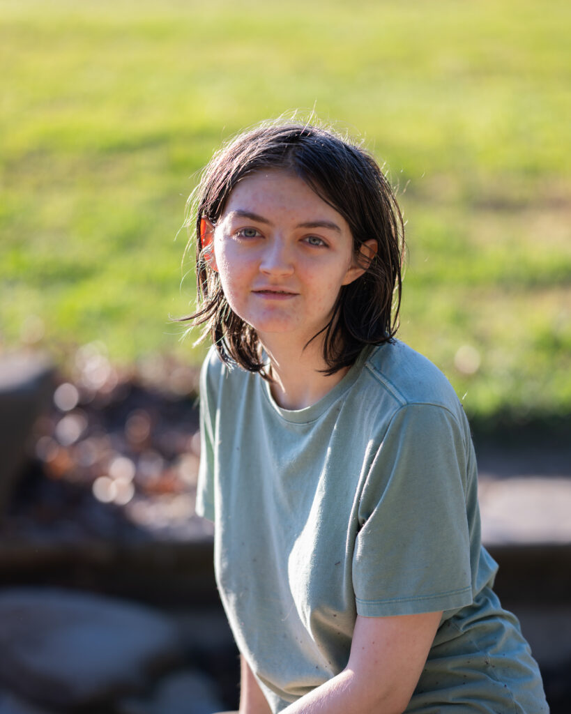 A Sacramento portrait photographer captures a young person with shoulder-length dark hair in a soft green T-shirt sitting outdoors. The background shows blurred greenery and warm golden light.