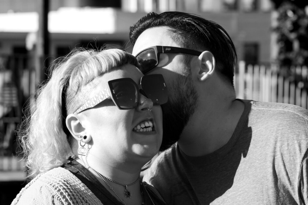 A black and white close up photo of couples photography. Both are wearing sunglasses and are in full sun. The man's face is partially obscured as he is kissing the side of her head. The woman is looking off to the side with a full teeth-bearing grin.