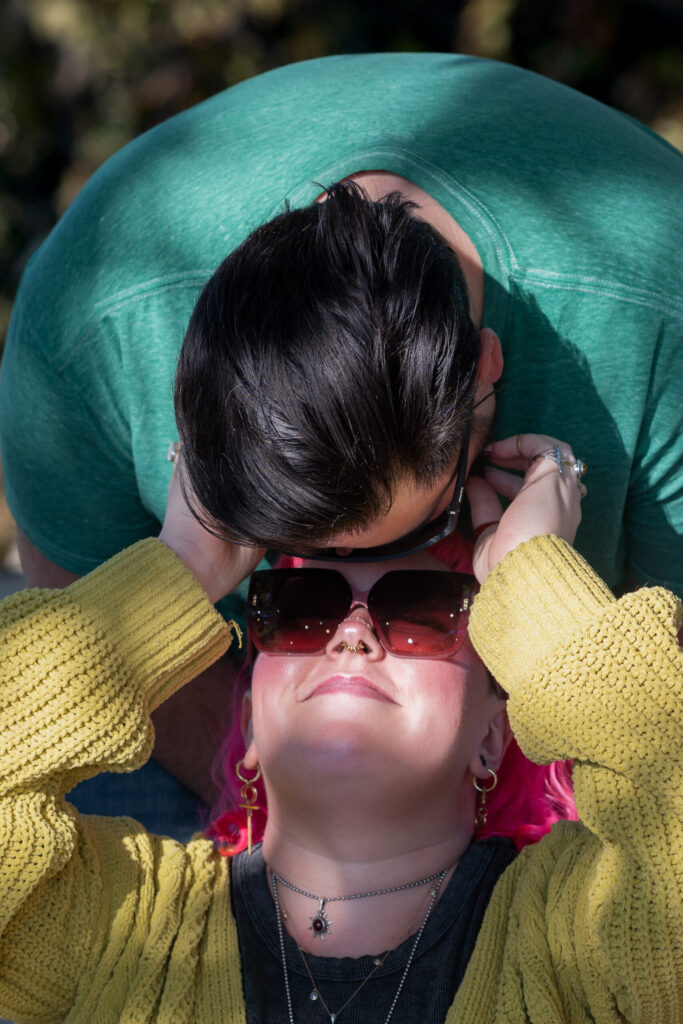 A man with black hair is leaning down over his girlfriend, kissing her forehead. She has bright pink hair and is wearing sunglasses, reaching up to scratch her boyfriend's beard.