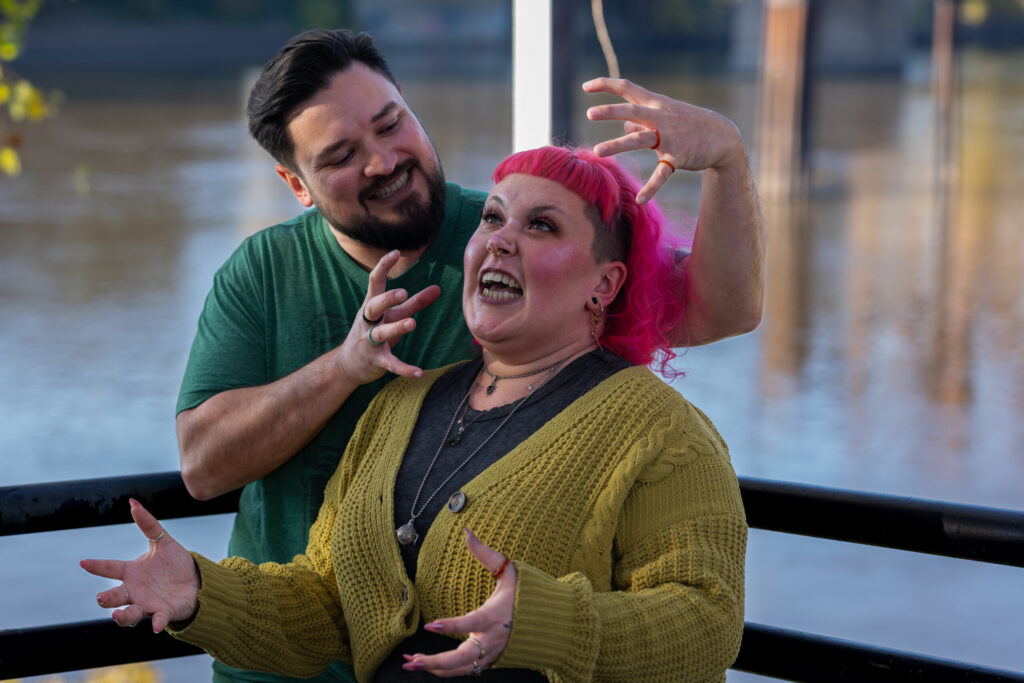 A couple leaning against a railing with water in the background. The woman has bright pink hair, has her hands out in front of her like claws with palms up, and is making a face. Her boyfriend is behind her and has black hair and a black beard, and has his hands in claws on either side of her head, smiling at her.