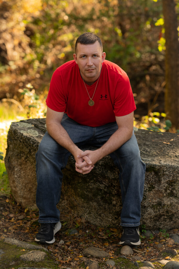 A Sacramento portrait photographer captures a man in a red shirt and jeans sitting on a large outdoor rock, looking at the camera. Warm sunlight filters through the trees.
